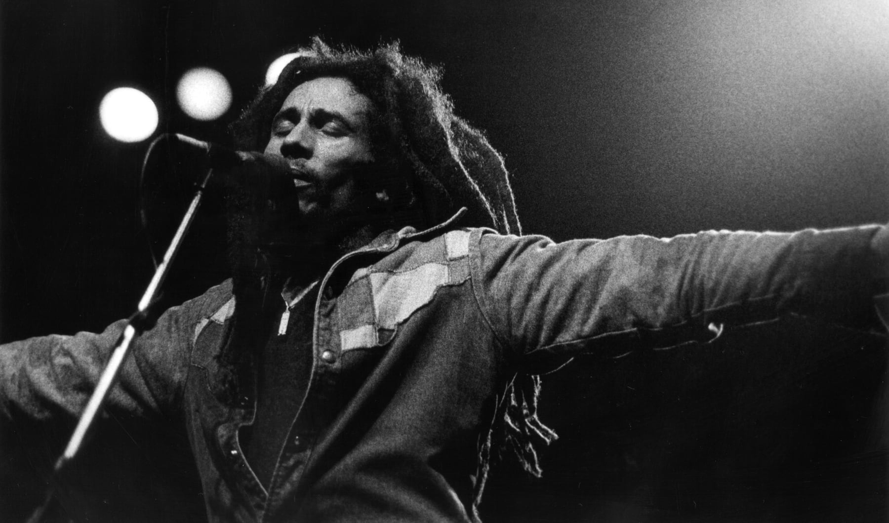 Bob Marley singing passionately on stage, arms stretched out, wearing a denim jacket, microphone in view.